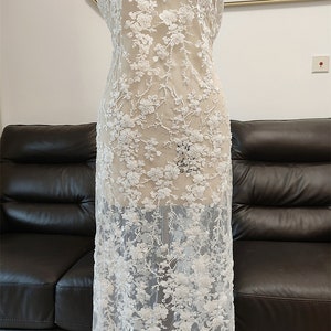 May include: White floral lace fabric with a delicate, embroidered design. The lace features a repeating pattern of flowers and leaves, creating a textured and elegant look. The fabric is displayed on a mannequin, showcasing its drape and detail.