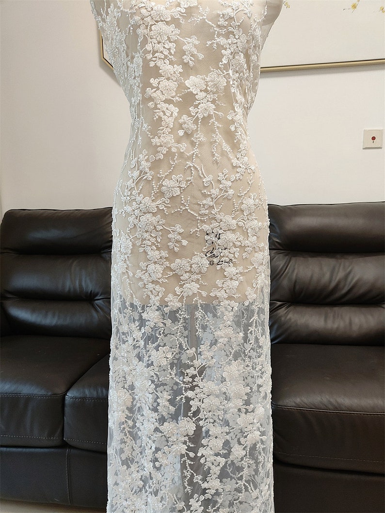 May include: White floral lace fabric with a delicate, embroidered design. The lace features a repeating pattern of flowers and leaves, creating a textured and elegant look. The fabric is displayed on a mannequin, showcasing its drape and detail.