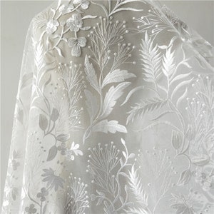 May include: White floral embroidered lace fabric with a delicate pattern of leaves and flowers. The lace is sheer and lightweight, making it perfect for a variety of projects.