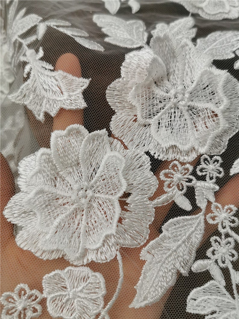 May include: White floral lace fabric with a delicate pattern of flowers and leaves. The lace is on a sheer mesh background.