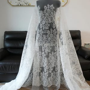 May include: White lace fabric with a floral pattern. The lace is draped over a mannequin, showcasing the intricate design. The fabric appears delicate and suitable for bridal or formal wear.