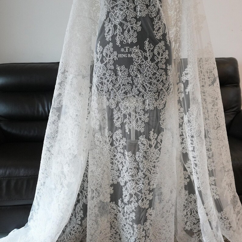 May include: White lace fabric with floral patterns. The delicate lace is draped over a form, showcasing the intricate design. The fabric is semi-transparent, allowing a glimpse of the form beneath. The text "FENG BA" is visible on the form.