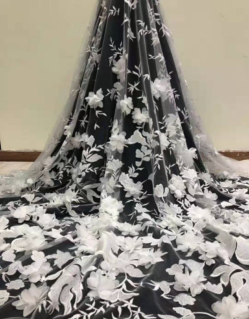 May include: White floral lace fabric with 3D flowers on a black background. The lace is delicate and intricate, with a soft, flowing texture. The flowers are large and prominent, adding a touch of elegance and sophistication to the fabric.