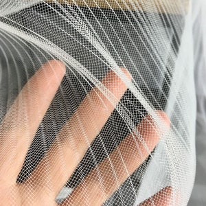 May include: White tulle fabric with a fine mesh pattern. The fabric is soft and delicate, and it has a slightly sheer appearance.