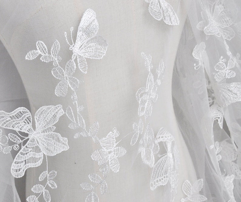 Pu&ograve; includere: Tessuto bianco trasparente con farfalle e motivi floreali ricamati in bianco.