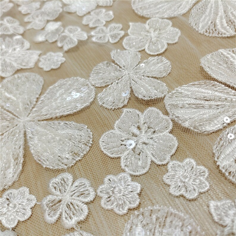 May include: White floral lace fabric with sequins. The fabric has a delicate, intricate design with small flowers and sequins. The fabric is sheer and lightweight, making it perfect for a variety of projects.