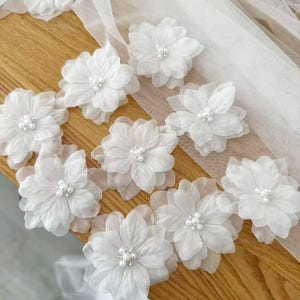 May include: A collection of white fabric flowers with layered petals and a cluster of small white faux pearls at the center of each bloom. The flowers are arranged in a scattered pattern, featuring soft, translucent material and rounded petal edges.