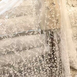 May include: White tulle fabric with a floral embroidery design. The embroidery is in a white color and features delicate leaves and small flowers. The fabric is sheer and has a soft, flowing texture.