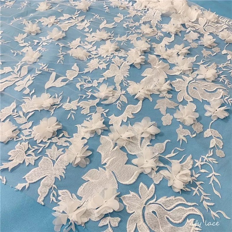 May include: White floral lace fabric with 3D flowers. The lace is on a light blue background. The fabric is delicate and intricate. The flowers are made of a soft, sheer material. The lace is perfect for wedding dresses, bridal gowns, and other special occasion garments.