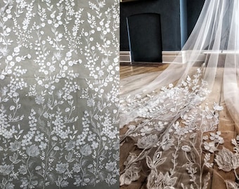 May include: White floral lace fabric with a delicate pattern of flowers and leaves. The lace is shown on a roll and laid out on a wooden floor.