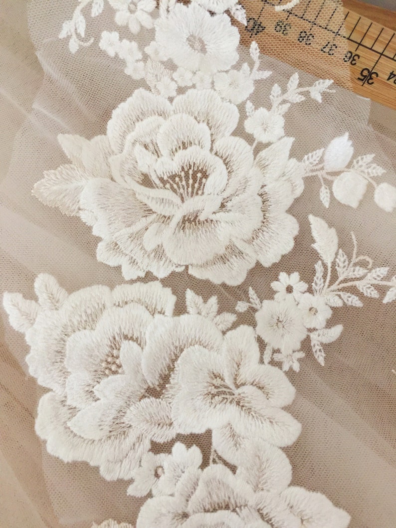 Puede incluir: Bordado floral blanco sobre una tela beige transparente. El bordado presenta flores grandes y peque&ntilde;as con detalles intrincados.