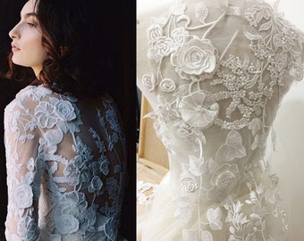 May include: A white lace wedding dress with a floral design. The dress has a long sleeve and a fitted bodice. The back of the dress features a large floral applique.
