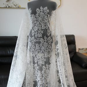 May include: White lace fabric with a floral pattern. The lace is draped over a mannequin, showcasing the intricate design. The fabric features a delicate, detailed floral motif, perfect for bridal or formal wear.