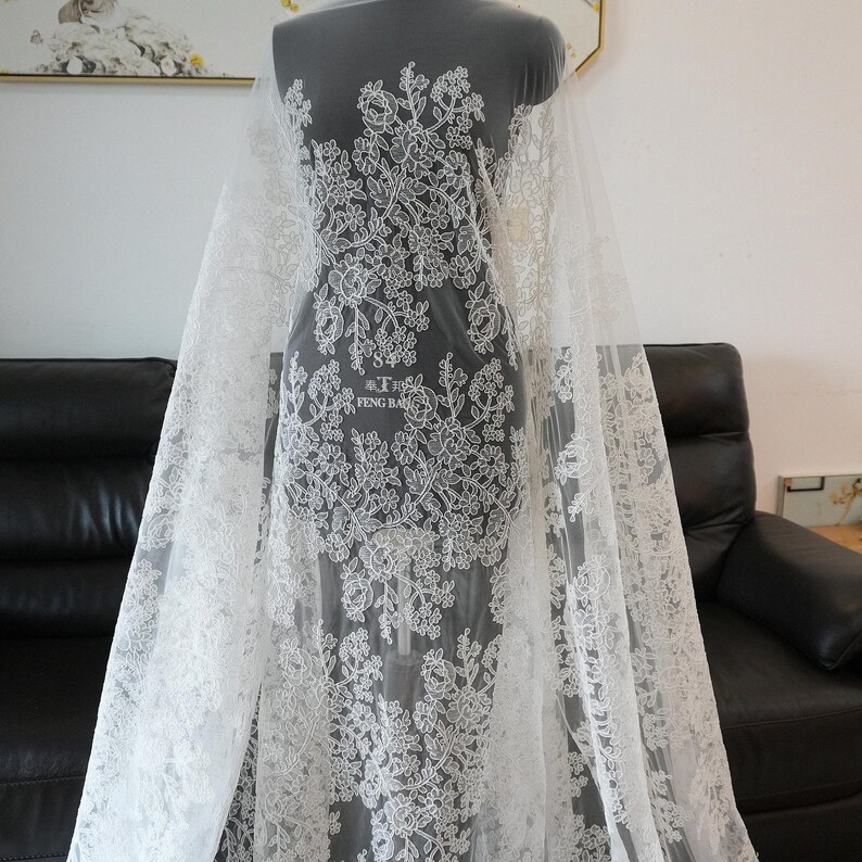 May include: White lace fabric with a floral pattern. The lace is draped over a mannequin, showcasing the intricate design. The fabric features a delicate, detailed floral motif, perfect for bridal or formal wear.