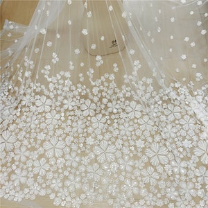 May include: White tulle fabric with white 3D floral embellishments. The fabric has a sheer, delicate texture and is adorned with intricate floral designs. The flowers are scattered throughout the fabric, creating a romantic and elegant look.