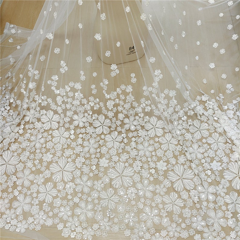 May include: White tulle fabric with white 3D floral embellishments. The fabric has a sheer, delicate texture and is adorned with intricate floral designs. The flowers are scattered throughout the fabric, creating a romantic and elegant look.