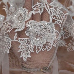 May include: White lace fabric with a floral pattern and sequins. The lace is delicate and intricate, with a soft, flowing texture.