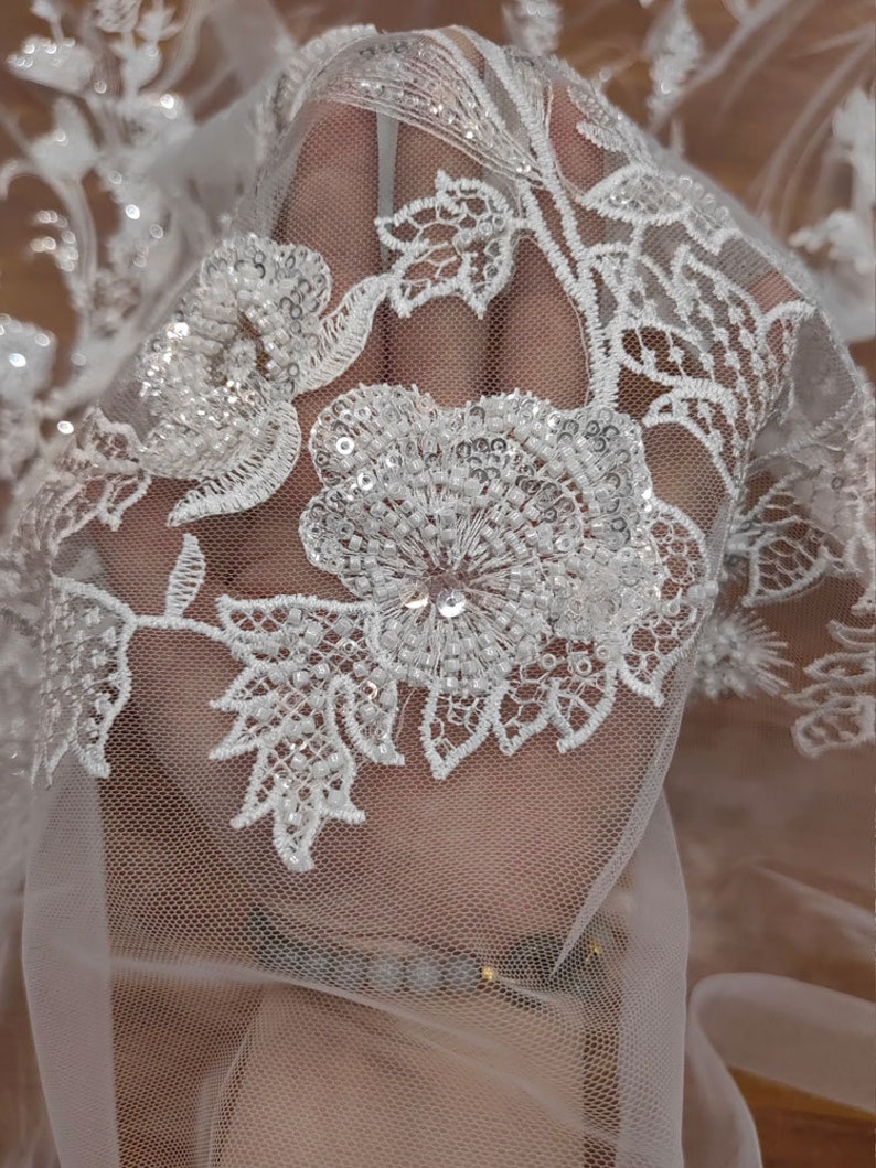 May include: White lace fabric with a floral pattern and sequins. The lace is delicate and intricate, with a soft, flowing texture.