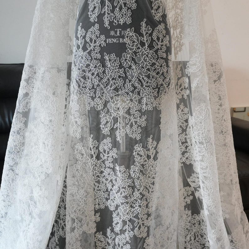May include: White lace fabric with floral patterns. The delicate lace features detailed embroidery of flowers and leaves. The fabric is draped over a form, showcasing the intricate design and texture. The text "奉.T.邦 FENG BA" is visible on the form.