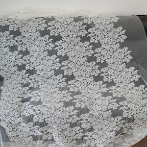 May include: White floral lace fabric with a delicate pattern of flowers and leaves. The lace is set against a gray background, creating a contrast that highlights the intricate design. This fabric is suitable for various sewing and crafting projects.
