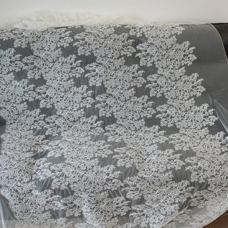 May include: White floral lace fabric with a delicate pattern of flowers and leaves. The lace is set against a gray background, creating a contrast that highlights the intricate design. This fabric is suitable for various sewing and crafting projects.