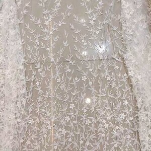 May include: White floral lace fabric with a delicate pattern of small white flowers and leaves. The lace is sheer and has a soft, romantic feel.