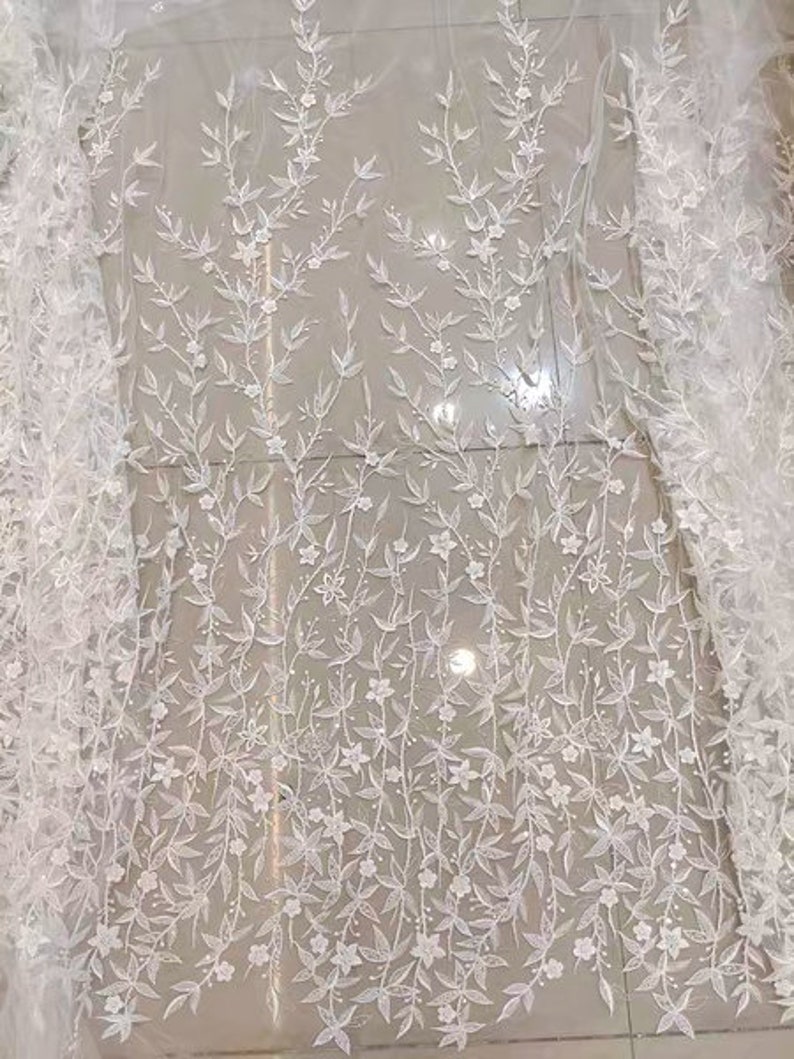 May include: White floral lace fabric with a delicate pattern of small white flowers and leaves. The lace is sheer and has a soft, romantic feel.