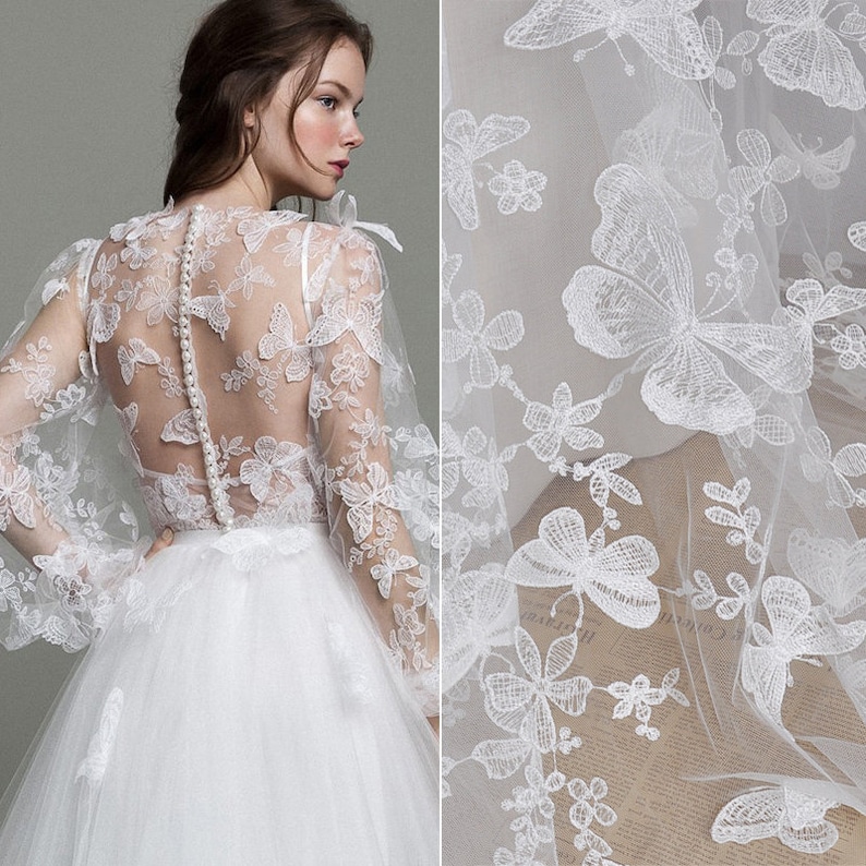 Pu&ograve; includere: Un abito da sposa bianco con un corpetto in pizzo trasparente con un motivo a farfalla. Il corpetto &egrave; abbottonato sul retro. La gonna &egrave; ampia e fluida.