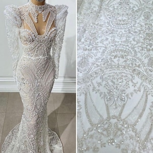 May include: A white, intricately beaded gown with long sleeves and a high neckline. The dress features a fitted bodice and a flowing skirt. The fabric on the right side of the image is a close-up of the same beaded material.
