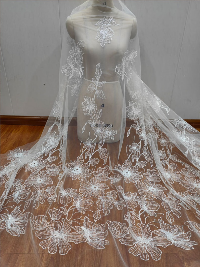 May include: White floral embroidered tulle fabric with a beaded design. The fabric is sheer and delicate, with a soft, flowing texture. The floral design is intricate and detailed, with small, white beads scattered throughout. The fabric is perfect for creating wedding gowns, bridesmaid dresses, and other special occasion garments.