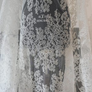 May include: White lace fabric with floral embroidery. The delicate design features roses and other flowers, creating a romantic and elegant look. The fabric is sheer, allowing light to pass through, and is suitable for bridal wear or other special occasion garments.