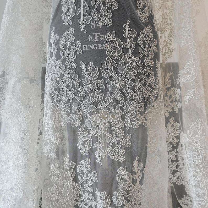 May include: White lace fabric with floral embroidery. The delicate design features roses and other flowers, creating a romantic and elegant look. The fabric is sheer, allowing light to pass through, and is suitable for bridal wear or other special occasion garments.