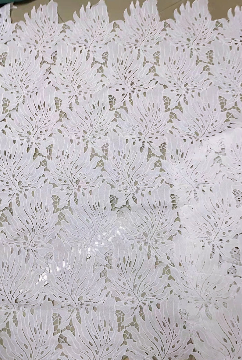 May include: White lace fabric with a delicate floral pattern. The fabric has a cut-out design that creates a see-through effect.