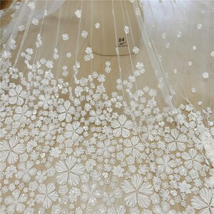 May include: White tulle fabric with white embroidered flowers. The flowers are 3D and have a shiny finish. The fabric has a sheer and delicate look. The fabric is labeled '84 cm'.
