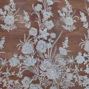 May include: White floral lace fabric with sequins. The fabric has a delicate floral pattern with sequins throughout. The fabric is on a brown wooden surface.
