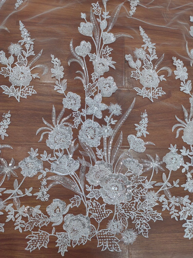 May include: White floral lace fabric with sequins. The fabric has a delicate floral pattern with sequins throughout. The fabric is on a brown wooden surface.