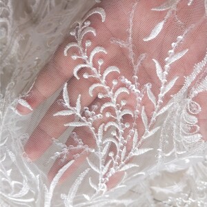 May include: White lace fabric with a floral pattern. The lace is delicate and has a soft, flowing texture. The fabric is perfect for wedding dresses, bridal veils, and other special occasion garments.