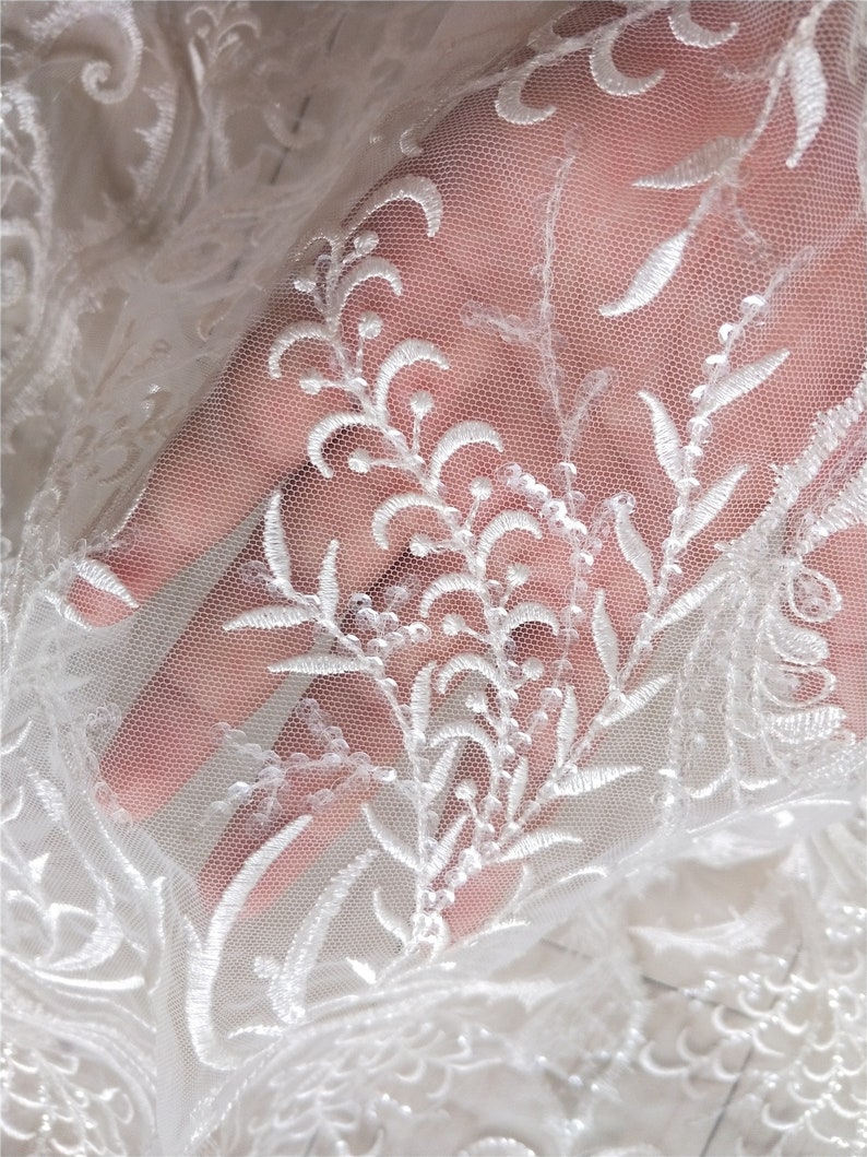 May include: White lace fabric with a floral pattern. The lace is delicate and has a soft, flowing texture. The fabric is perfect for wedding dresses, bridal veils, and other special occasion garments.
