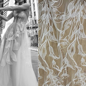 May include: Close-up view of white embroidered tulle fabric featuring a floral design. The embroidery is detailed and delicate, with a repeating pattern of stylized flowers and leaves. The tulle is sheer and lightweight, allowing the underlying surface to show through. The fabric is ideal for wedding dresses, bridal gowns, or other special occasion garments. The design is elegant and sophisticated, with a romantic feel.