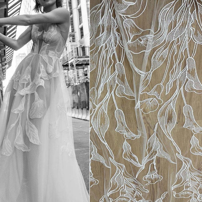 May include: Close-up view of white embroidered tulle fabric featuring a floral design. The embroidery is detailed and delicate, with a repeating pattern of stylized flowers and leaves. The tulle is sheer and lightweight, allowing the underlying surface to show through. The fabric is ideal for wedding dresses, bridal gowns, or other special occasion garments. The design is elegant and sophisticated, with a romantic feel.