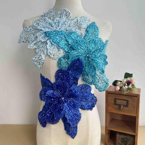 Może przedstawiać: Trzy niebieskie i białe aplikacje kwiatowe wykonane z cekinów i koronki. Aplikacje są ułożone jedna na drugiej i ułożone w kaskadowym wzorze. Aplikacje są idealne do dodania odrobiny elegancji do każdego ubrania lub projektu.