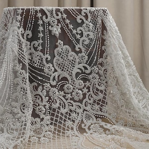 May include: White beaded lace fabric featuring a floral design. The fabric is delicate and intricate, with small pearls sewn throughout.