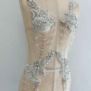 May include: A white bodice appliqué set featuring intricate silver embroidery, clear faceted crystals, and delicate seed beads. The design includes two upper and two lower floral-shaped sections connected by multiple draped strands of clear beads.
