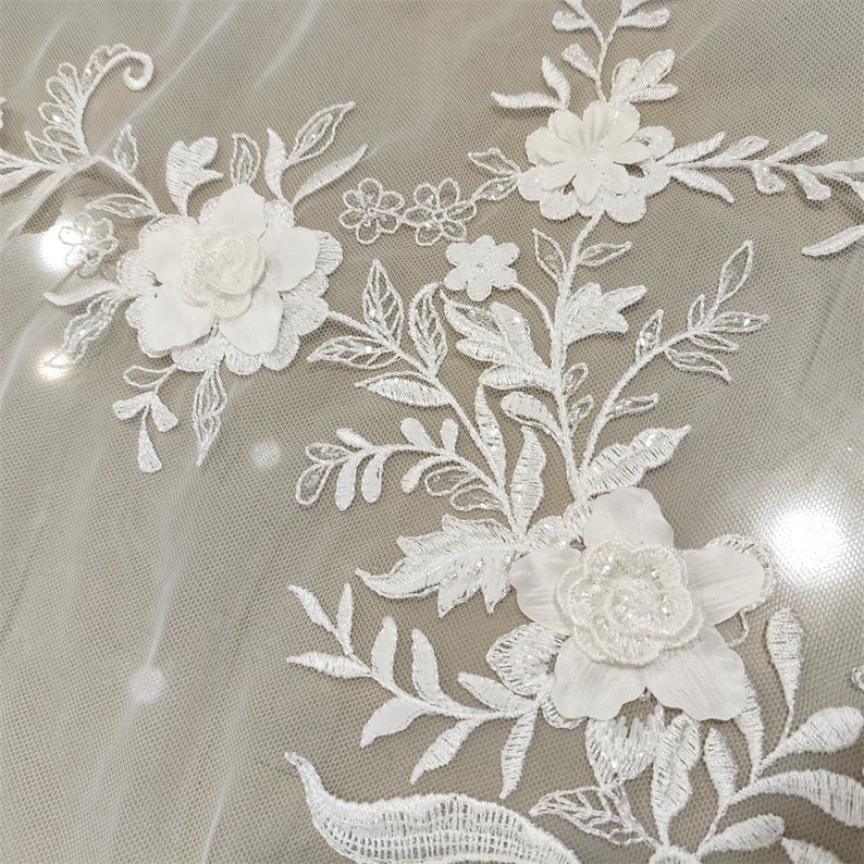 May include: White floral lace fabric with 3D flowers and sequins. The fabric has a delicate and intricate design with a sheer background.