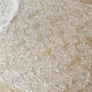 Peut inclure: Tissu de dentelle florale blanche avec un motif délicat et complexe. La dentelle est sur un fond beige et a une texture douce et fluide.