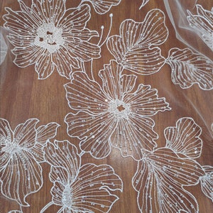 May include: White floral lace fabric with a beaded design. The fabric has a delicate, intricate pattern of flowers and leaves. The beads are small and evenly spaced, creating a subtle shimmer.