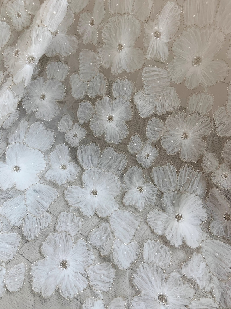 May include: White floral lace fabric with 3D flowers and pearl embellishments. The fabric has a delicate and intricate design.