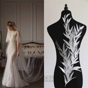 May include: A white lace applique featuring a delicate floral design with a leafy pattern. The applique is shown on a black mannequin and is perfect for adding a touch of elegance to a wedding dress or other special occasion garment.