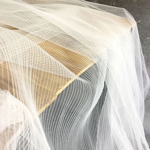 May include: White sheer fabric with a striped pattern. The fabric is draped over a wooden surface.