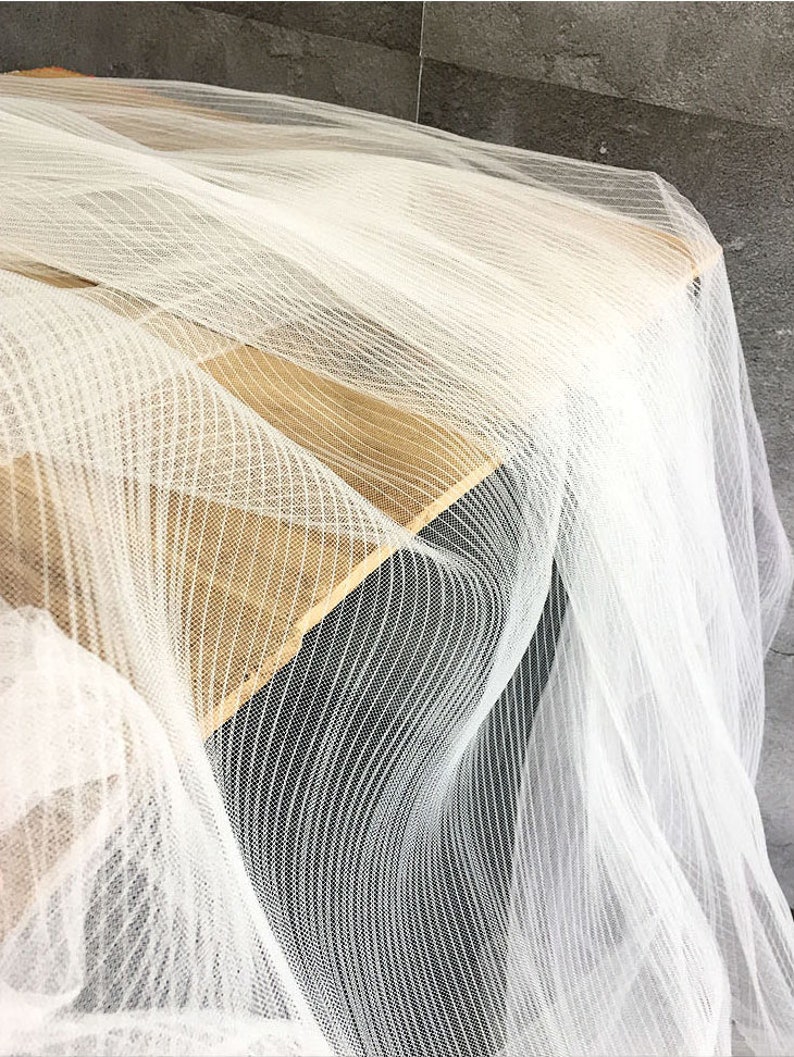 May include: White sheer fabric with a striped pattern. The fabric is draped over a wooden surface.